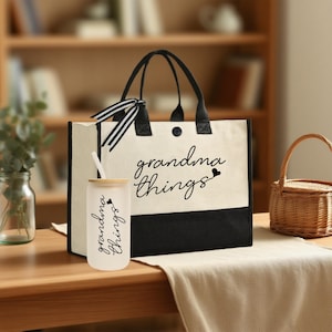Peut inclure: Un sac fourre-tout en toile crème avec des poignées noires et une base noire, et un gobelet en verre, tous deux avec l'inscription "grandma things" en écriture noire. Le gobelet a un couvercle en bambou et une paille.