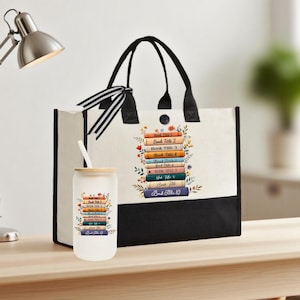 May include: A tote bag and a glass tumbler with a book theme. The tote bag is white with black accents and handles, featuring a stack of books with floral accents. The tumbler has a straw and a matching book design.
