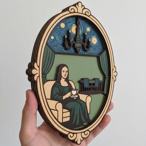May include: A layered wooden art piece featuring a reimagining of the Mona Lisa. The artwork depicts the Mona Lisa seated in a room, holding a teacup, with a starry night sky visible through a window. The frame is oval-shaped.