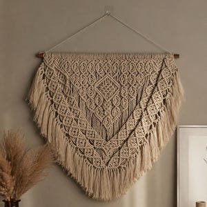 May include: Beige macrame wall hanging with a geometric diamond pattern and fringe. The wall decor is suspended from a wooden dowel and a string. This handcrafted piece adds a bohemian touch to any room.