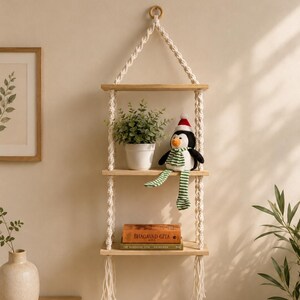 May include: A three-tier hanging wall shelf made of light-coloured wood planks suspended by thick, cream-coloured macrame rope. The shelves hold a small potted green plant, a plush penguin wearing a Santa hat and scarf, and a book titled Bhagavad-Gita.