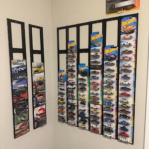 May include: Black wall-mounted display shelves filled with various Hot Wheels toy cars in their original packaging. The collection includes a variety of car models and colors, arranged neatly on the shelves. A Matchbox Convoy toy is also displayed.