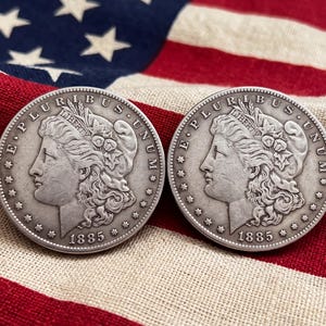 Puede incluir: Dos monedas de dólar Morgan de tono plateado fechadas en 1885, cada una con el perfil de Lady Liberty, el texto E PLURIBUS UNUM y trece estrellas a lo largo del borde. Las monedas son circulares con acabado metálico y grabados en relieve.