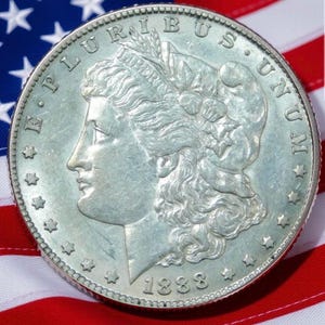 Pode incluir: Uma moeda de prata Morgan de 1888, com um perfil de Lady Liberty e as palavras "E PLURIBUS UNUM". A moeda est&aacute; colocada sobre um fundo da bandeira americana, com as suas riscas vermelhas e brancas e o seu campo azul de estrelas.