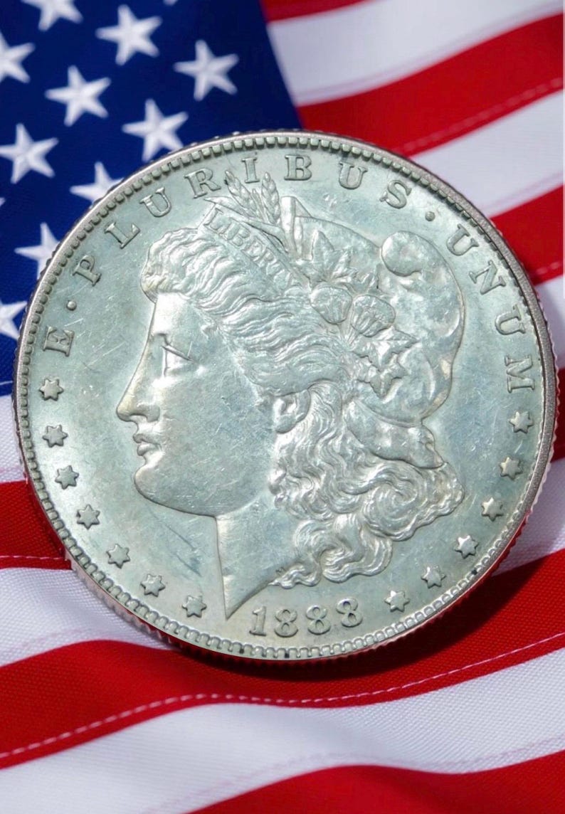 Pode incluir: Uma moeda de prata Morgan de 1888, com um perfil de Lady Liberty e as palavras "E PLURIBUS UNUM". A moeda est&aacute; colocada sobre um fundo da bandeira americana, com as suas riscas vermelhas e brancas e o seu campo azul de estrelas.