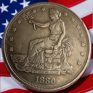 Puede incluir: Primer plano de una moneda de oro antigua de 1880, con una mujer sentada sosteniendo una rama y trigo. La moneda está colocada frente a la bandera estadounidense, con sus estrellas y rayas visibles.