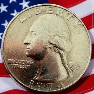 Może przedstawiać: Zbliżenie monety ćwierćdolarowej z 1974 roku z profilem George'a Washingtona, napisami "LIBERTY" i "IN GOD WE TRUST". Moneta jest umieszczona na tle amerykańskiej flagi z czerwono-białymi pasami i niebieskim polem z gwiazdami.