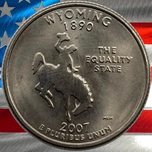 Może przedstawiać: Srebrna moneta ćwierćdolarowa stanu Wyoming z kowbojem na koniu. Moneta zawiera napisy "WYOMING 1890" i "THE EQUALITY STATE". Moneta jest datowana na 2007 rok i ma napis "E PLURIBUS UNUM". Tło to flaga amerykańska.