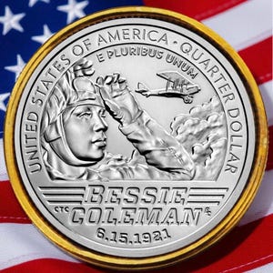 May include: A silver quarter dollar coin with a gold rim, featuring a portrait of Bessie Coleman and an airplane. The coin is set against a backdrop of the American flag, with red and white stripes and blue stars.
