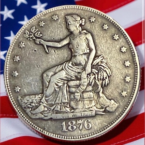 May include: A silver coin featuring a seated woman holding a sprig, surrounded by stars. The coin is dated 1876 and the words "IN GOD WE TRUST" are visible. The coin is set against a backdrop of the American flag.