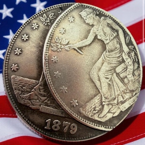 Puede incluir: Dos monedas de plata con grabados detallados, que representan a una mujer y el año 1879, descansan sobre una bandera estadounidense. Las monedas tienen diseños y texto intrincados, con un campo azul estrellado y rayas rojas y blancas.