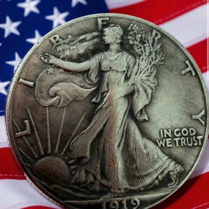 May include: A silver Walking Liberty half dollar coin from 1919, featuring Lady Liberty walking towards the sun, with the words "In God We Trust." The coin is set against a backdrop of the American flag.
