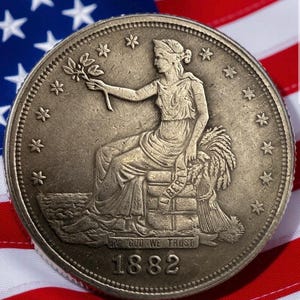 May include: A detailed close-up of an antique coin with a seated woman holding a branch, surrounded by stars. The coin is set against a backdrop of the American flag. The year 1882 is visible on the coin.