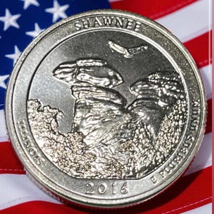 May include: A silver quarter coin featuring a detailed relief of rock formations, with the inscription "SHAWNEE" and "ILLINOIS". The coin is dated 2016 and set against a backdrop of the American flag.