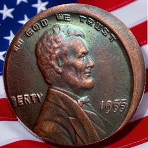 May include: A close-up of a 1955 Lincoln penny with the words "In God We Trust" and "Liberty" visible. The penny is copper-colored and set against a backdrop of the American flag.