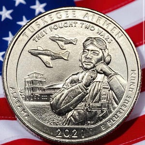 Pode incluir: Uma moeda de prata com um retrato de um aviador de Tuskegee, dois aviões de combate e a inscrição "Tuskegee Airmen They Fought Two Wars". A moeda está colocada sobre o fundo da bandeira americana, com o ano de 2021 visível.