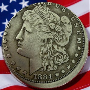 May include: A silver 1884 Morgan dollar coin with a profile of Liberty, surrounded by stars and the inscription "LIBERTY" and "UNITED STATES OF AMERICA". The coin is set against a backdrop of the American flag.