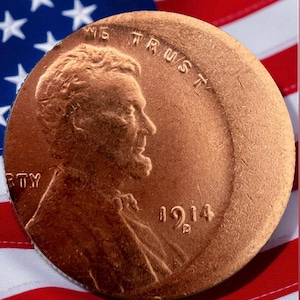 Peut inclure: Gros plan d'un penny Lincoln de 1914 de couleur cuivre, avec les inscriptions « In God We Trust » et le chiffre « 1914 ». La pièce est posée sur un fond de drapeau américain, avec des rayures rouges et blanches et un champ d'étoiles bleu.