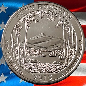 May include: A silver quarter coin featuring a detailed landscape of White Mountain, New Hampshire, with trees, a lake, and mountains. The coin is inscribed with "White Mountain", "New Hampshire", "2013", and "E Pluribus Unum". The background shows the American flag.