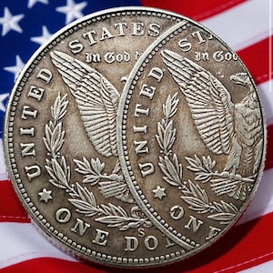 May include: Two silver dollar coins with detailed engravings of an eagle and the words "United States of America" and "One Dollar." The coins are set against a backdrop of the American flag, with its red and white stripes and blue field of stars.
