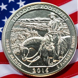 May include: A silver coin featuring Theodore Roosevelt on horseback, with the text "THEODORE ROOSEVELT" and "NORTH DAKOTA" around the edge. The coin is dated 2016 and rests on an American flag.