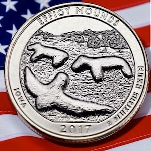 May include: A silver quarter coin featuring the Effigy Mounds National Monument in Iowa. The coin displays three mound shapes, the text "EFFIGY MOUNDS", "IOWA", "2017", and "E PLURIBUS UNUM". The coin is set against a backdrop of the American flag.