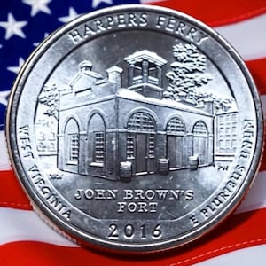 May include: A silver quarter coin featuring John Brown's Fort in Harpers Ferry, West Virginia, with the year 2016. The coin is set against a backdrop of the American flag, with red and white stripes and stars.