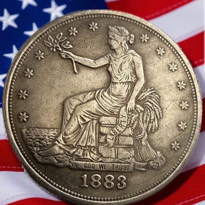 Puede incluir: Primer plano de una moneda antigua con una figura femenina sentada sosteniendo una ramita. La moneda, fechada en 1883, presenta la inscripción "IN GOD WE TRUST" y la palabra "LIBERTY". La moneda está sobre un fondo de la bandera estadounidense.