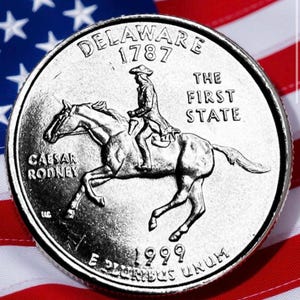 May include: A silver Delaware quarter coin from 1999, featuring Caesar Rodney on horseback, with the text "DELAWARE 1787" and "THE FIRST STATE". The coin is set against a backdrop of the American flag.