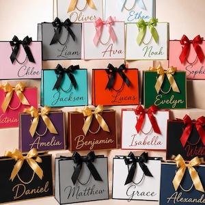 May include: A collection of rectangular gift bags in various colours, each featuring a satin ribbon bow, gold-tone cord handles, and a personalised name printed in a cursive script. The bags are arranged in a tiered display, showcasing diverse colour combinations.