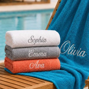 May include: A stack of three folded towels in white, gray, and orange, each embroidered with a name: Sophia, Emma, and Ava. A large blue towel is draped over a wooden lounge chair, featuring the embroidered name Olivia in white script.