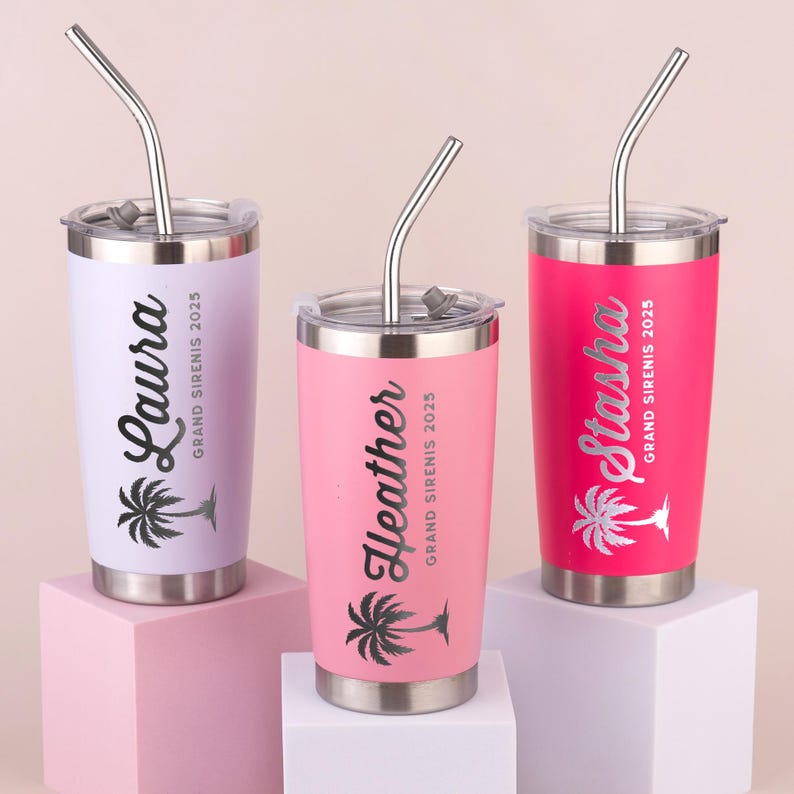 May include: Three stainless steel tumblers in pastel colors: lavender, pink, and hot pink. Each tumbler has a silver lid and straw. The names "Laura", "Heather", and "Stasha" are printed on the tumblers, along with a palm tree graphic and the text "GRAND SIRENIS 2025".