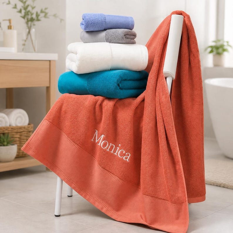 May include: A stack of folded towels in light blue, gray, white, and teal sits atop a large, draped, burnt orange towel. The orange towel features the embroidered name Monica in white script lettering along its wide, smooth bottom border.