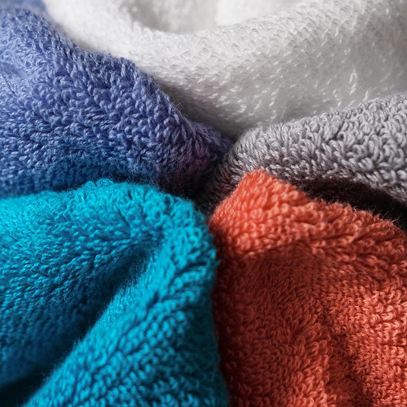 May include: A collection of folded terry cloth towels in a variety of colors including white, light gray, deep blue, bright turquoise, and burnt orange. The fabric features a dense, looped texture characteristic of absorbent cotton bath towels.