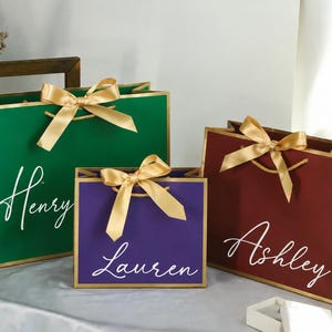 May include: Three rectangular gift bags in green, purple, and maroon, each featuring gold trim, a gold ribbon bow, and white cursive text reading Henry, Lauren, and Ashley respectively. The bags have gold cord handles and a smooth, matte finish.