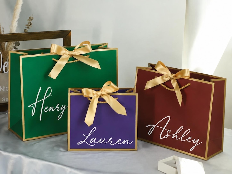 May include: Three rectangular gift bags in green, purple, and maroon, each featuring gold trim, a gold ribbon bow, and white cursive text reading Henry, Lauren, and Ashley respectively. The bags have gold cord handles and a smooth, matte finish.