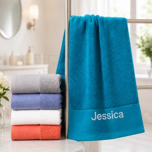 May include: A stack of four folded cotton towels in gray, light blue, white, and coral, positioned next to a hanging teal towel featuring the embroidered name Jessica in a light gray script font on the lower border of the fabric.