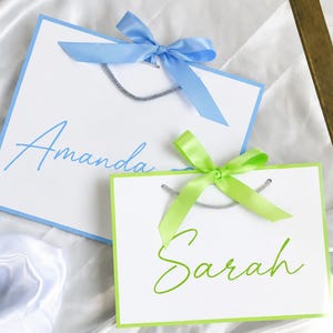 May include: Two white gift bags with silver cord handles. The larger bag features a light blue border, a light blue satin ribbon bow, and the name Amanda in blue script. The smaller bag has a lime green border, a lime green satin ribbon bow, and the name Sarah in lime green script.