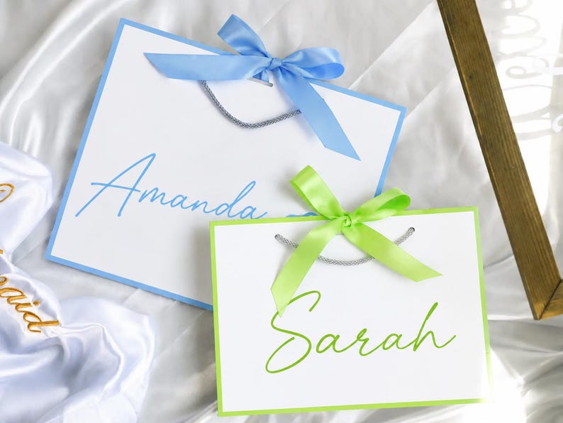 May include: Two white gift bags with silver cord handles. The larger bag features a light blue border, a light blue satin ribbon bow, and the name Amanda in blue script. The smaller bag has a lime green border, a lime green satin ribbon bow, and the name Sarah in lime green script.