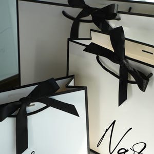 May include: Three white gift bags with black trim and black ribbon handles tied in bows. The bags feature black script text on the front, with the names Lauren and Natalia clearly visible. The bags are arranged in a cluster, showing different sizes.
