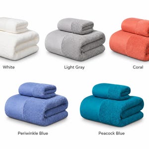 May include: Five sets of folded towels, each consisting of one large bath towel and one smaller hand towel. The sets are available in five distinct solid colors: white, light gray, coral, periwinkle blue, and peacock blue, all featuring a textured border.