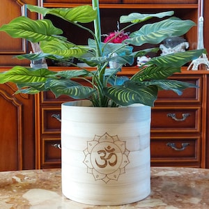 Peut inclure: Un pot de fleurs cylindrique de couleur claire avec un symbole Om sculpté, contenant une plante verte feuillue. Le pot est posé sur une surface à motifs de marbre. Un seau en cuivre et un bougeoir décoratif sont également visibles.