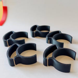May include: Five black, C-shaped plastic clips with a textured finish. The clips are arranged on a light background. Each clip has a curved top and a flat base, with a small gap in the middle. The design is simple and functional.