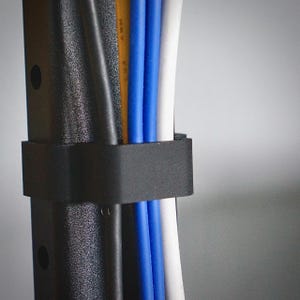 May include: Close-up of a black cable organizer securing a bundle of colorful wires. The wires include black, white, blue, and yellow cables. The organizer is made of a textured material.