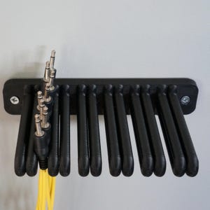 Op de afbeelding: Een zwarte wandhouder voor kabels met meerdere sleuven voor het opbergen van audiokabels. De houder is bevestigd met twee zilverkleurige schroeven en bevat diverse zwarte audiokabels met zilverkleurige 6,35 mm jackpluggen en felgele snoeren.