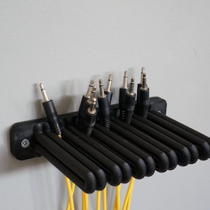Peut inclure: Un support mural noir pour câbles doté de plusieurs fentes conçues pour maintenir des câbles audio. Le support est fixé avec des vis visibles et contient plusieurs prises audio noires avec des embouts métalliques, avec des câbles jaunes.