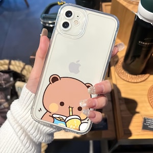 May include: Clear phone case with a cartoon bear design. The bear is light brown and is drinking from a yellow cup with a blue and white design. The case has a clear border and is designed for an iPhone.