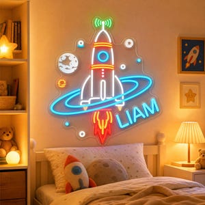 May include: A wall-mounted neon sign featuring a rocket ship design with a blue ring, a green antenna, and red flames. The sign includes the name LIAM in blue neon letters, surrounded by small circular planet and star accents on a clear backing.