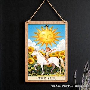 Könnte beinhalten: Holzschild mit einer Tarotkarten-Illustration der Sonne. Ein Kind reitet auf einem weißen Pferd durch Sonnenblumen unter einer lächelnden Sonne. Der Text "THE SUN" befindet sich unten.