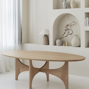 Puede incluir: Mesa de comedor ovalada de madera clara. La mesa tiene una superficie lisa y una base única con patas curvas. La habitación tiene una paleta de colores neutros con elementos decorativos en nichos.